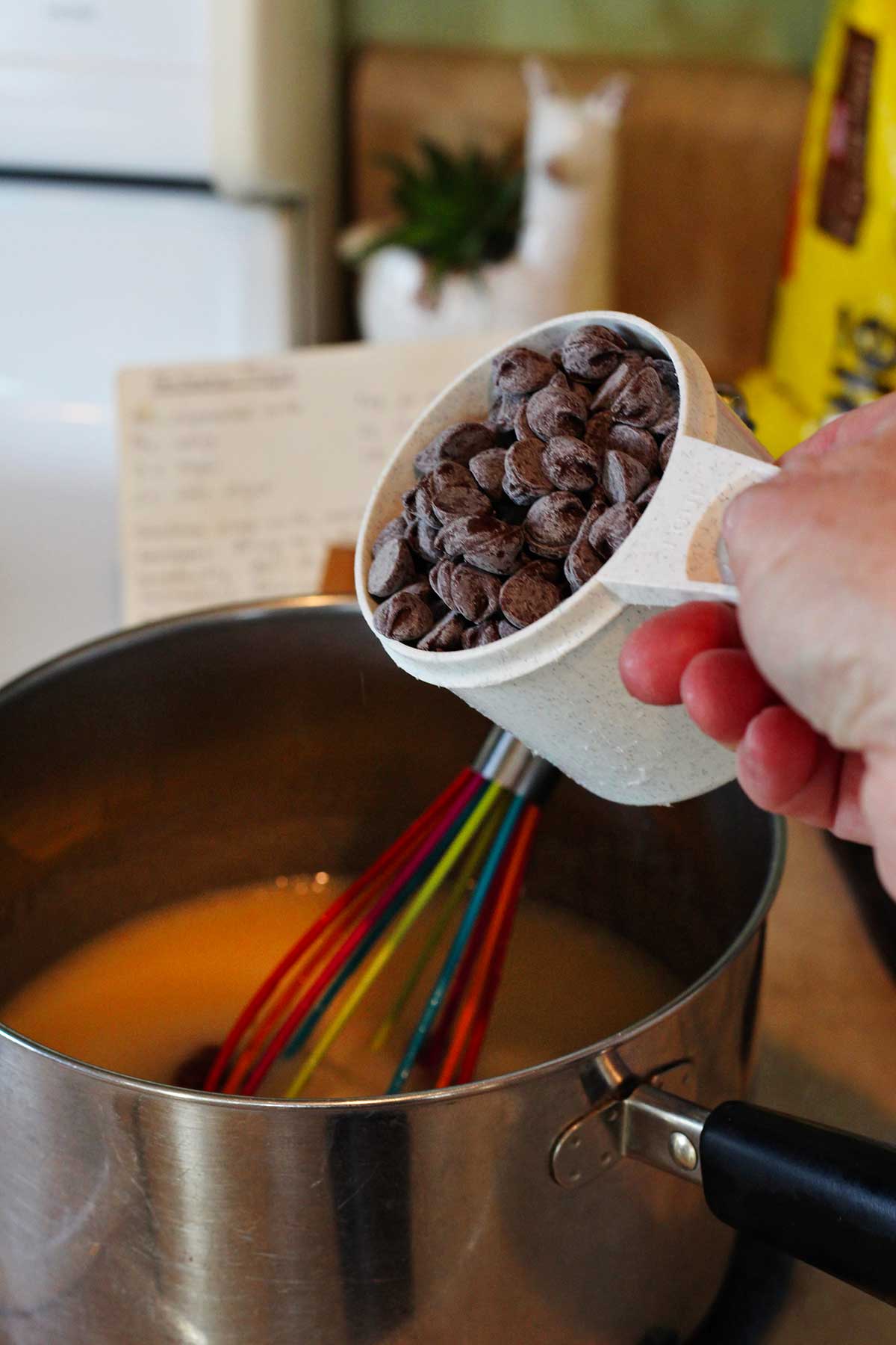 A person about to add a cup of chocolate chips to fudge mixture in saucepan.