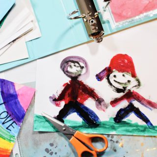 A child's painting of two people holding hands, with scissors, a binder and scrapbook paper nearby.