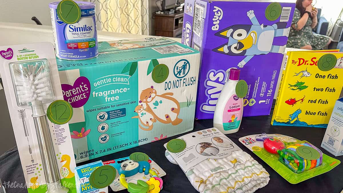 Baby shower game setup with baby items labeled by number, including diapers, wipes, baby lotion, formula, book, toys, and bottle brush for a Price Is Right guessing game.