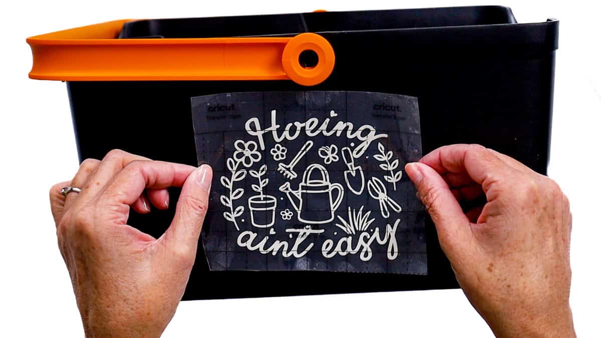 Hands position a gardening-themed vinyl decal with transfer tape onto the front of a black garden bucket before pressing it in place.