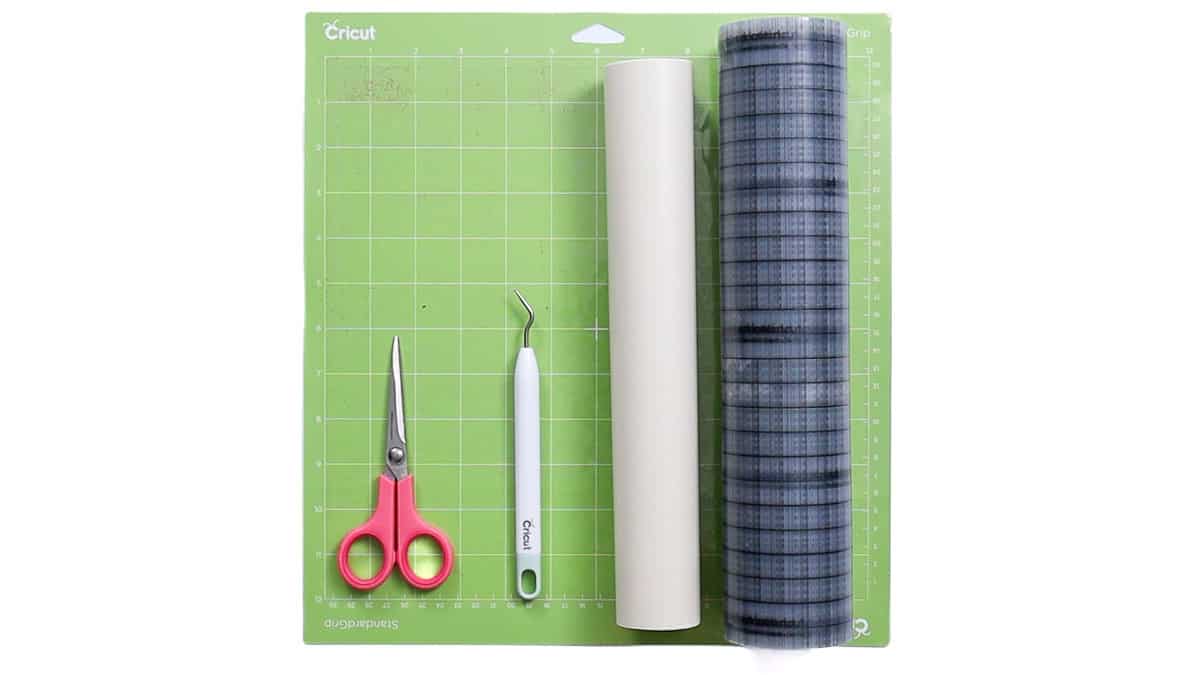 Cricut cutting mat with scissors, weeding tool, roll of adhesive vinyl, and transfer tape arranged neatly on top.