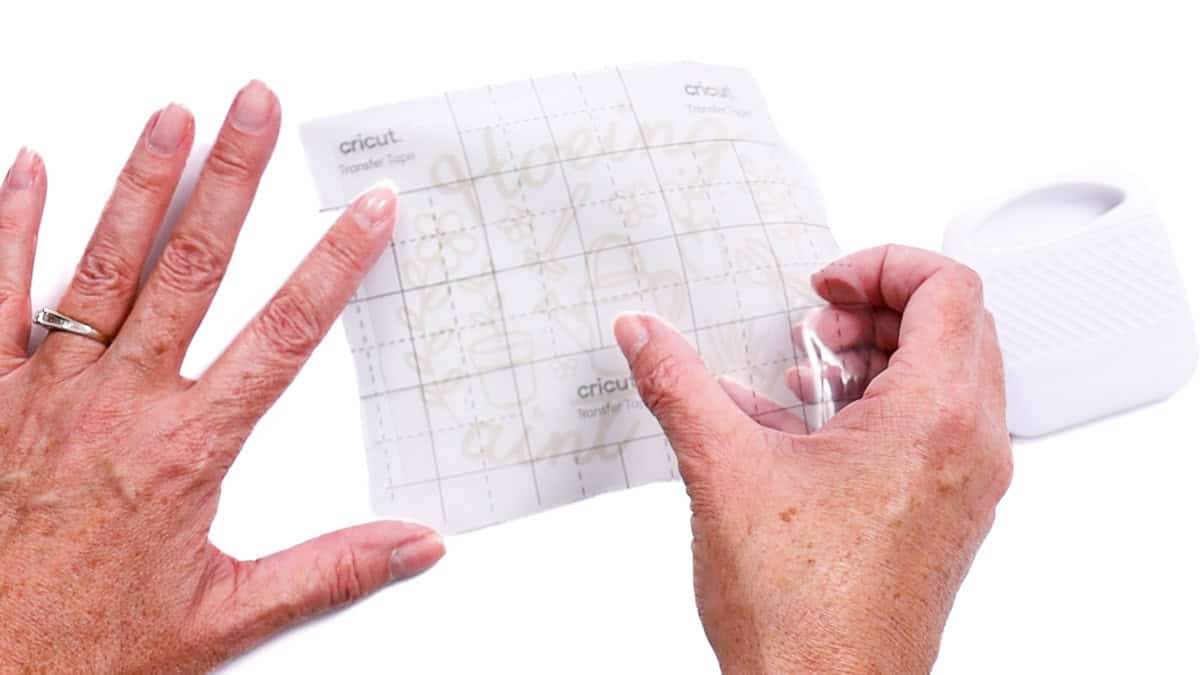 Hands apply transfer tape over a weeded vinyl decal, smoothing it down with a scraper tool to prepare for application.