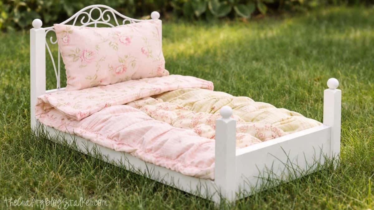 Ruffled Baby Doll Quilt Tutorial Handmade ruffled quilt and pillow on a small white doll bed placed on grass, showing a finished doll bedding set outdoors.