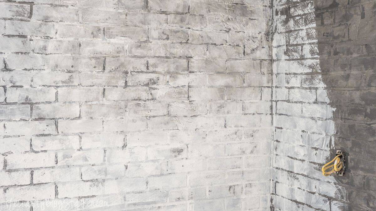 Faux brick wall partially whitewashed, showing layered paint over brick texture with visible mortar lines and a corner section still in progress.