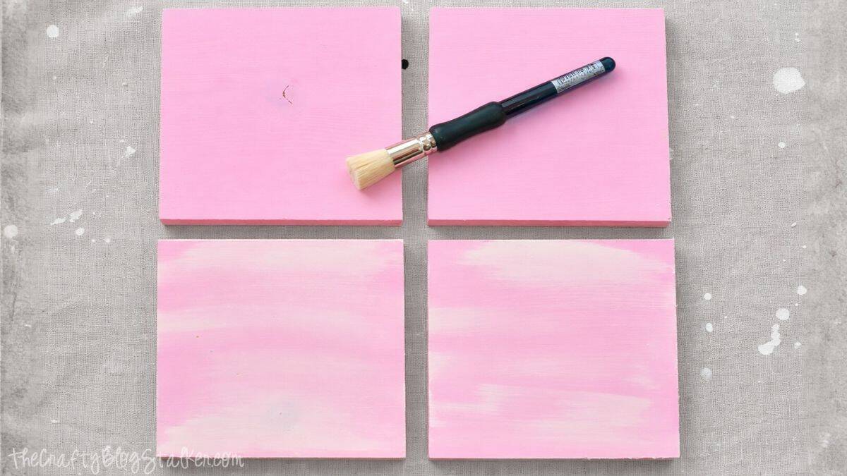 Four square wooden boards painted and laid flat on a work surface, with a paintbrush resting across the top boards during a DIY painting project.