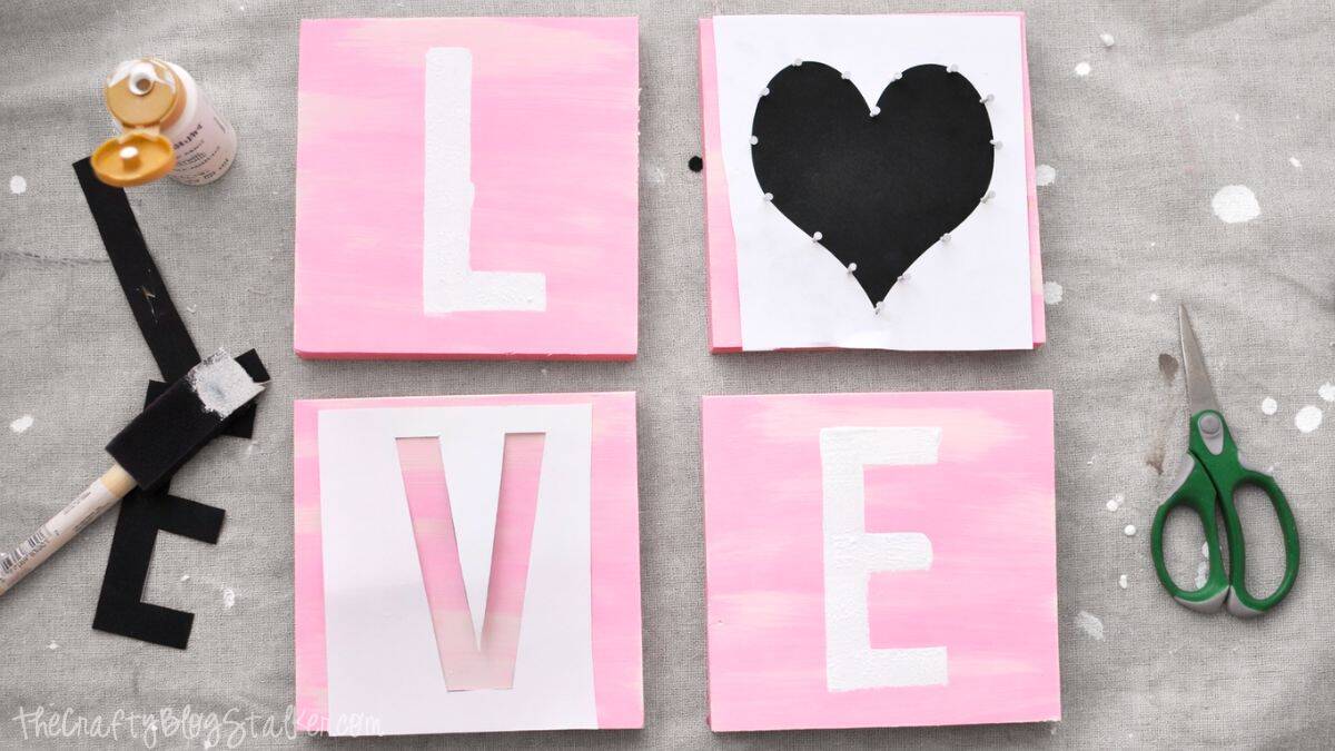 Four painted wooden squares arranged to spell LOVE, with letter stencils, a heart template, paint, brush, tape, and scissors laid out during a DIY banner project.