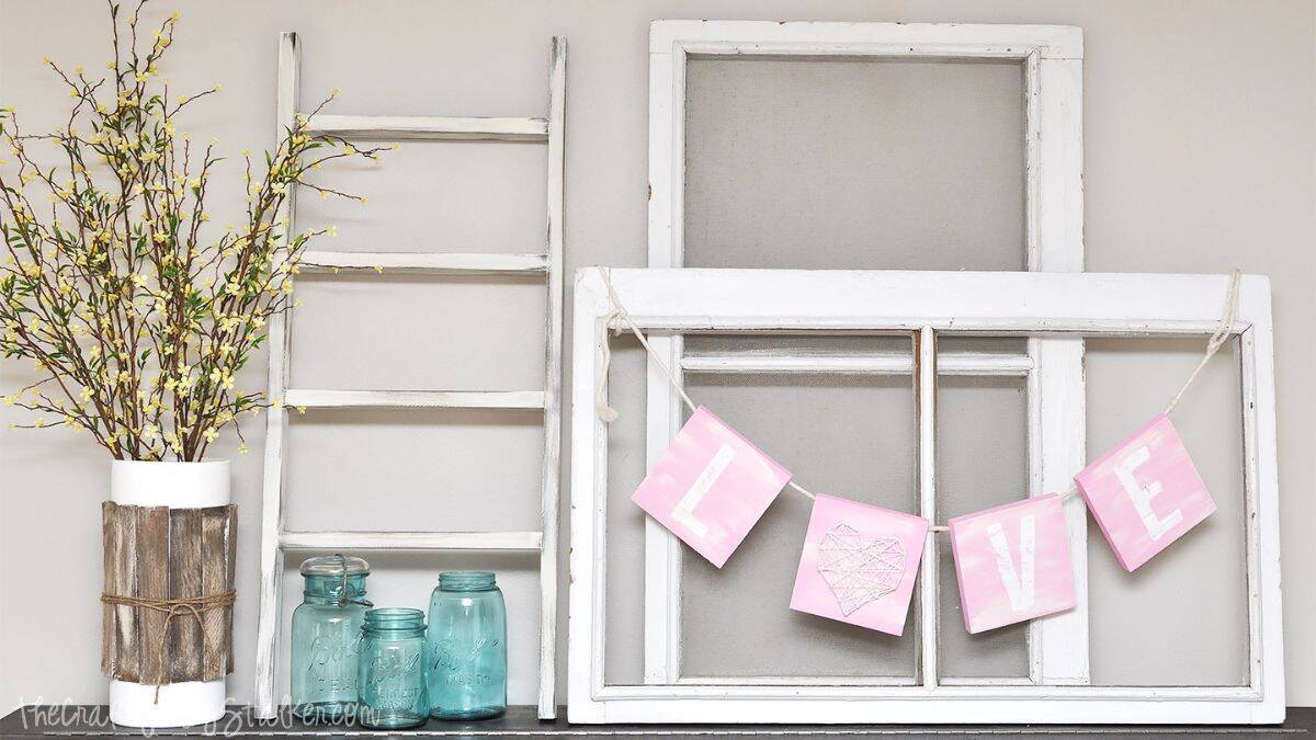 Valentine’s mantel decor featuring a DIY LOVE banner hung on a vintage window frame, with a rustic ladder, glass jars, and floral arrangement.