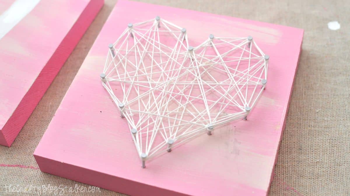 Heart-shaped string art made with nails and wrapped string on a painted wooden square, shown as part of a DIY Valentine decor project.