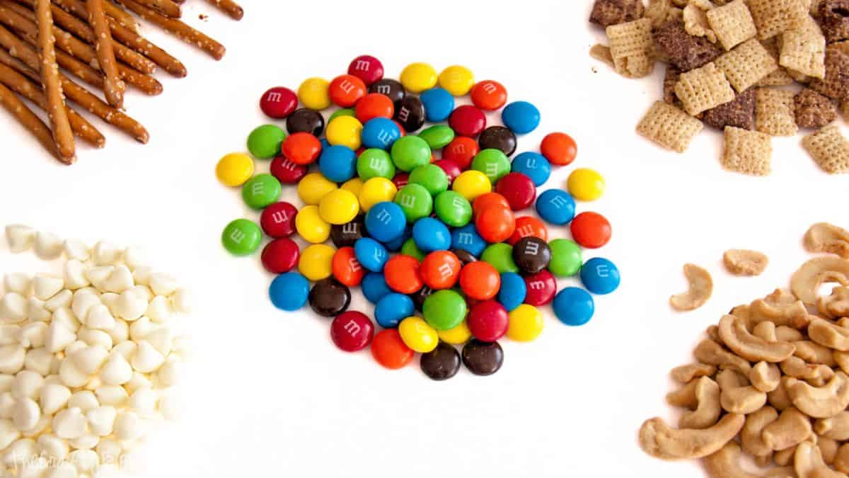 M&M White Chocolate Party Mix Recipe Ingredients for white chocolate party mix arranged on a white surface, including pretzels, cereal squares, white chocolate chips, cashews, and candy-coated chocolates.