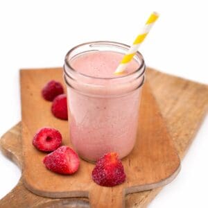 Strawberry smoothie in a mason jar with a striped paper straw, placed on a wooden cutting board with frozen strawberries beside it.