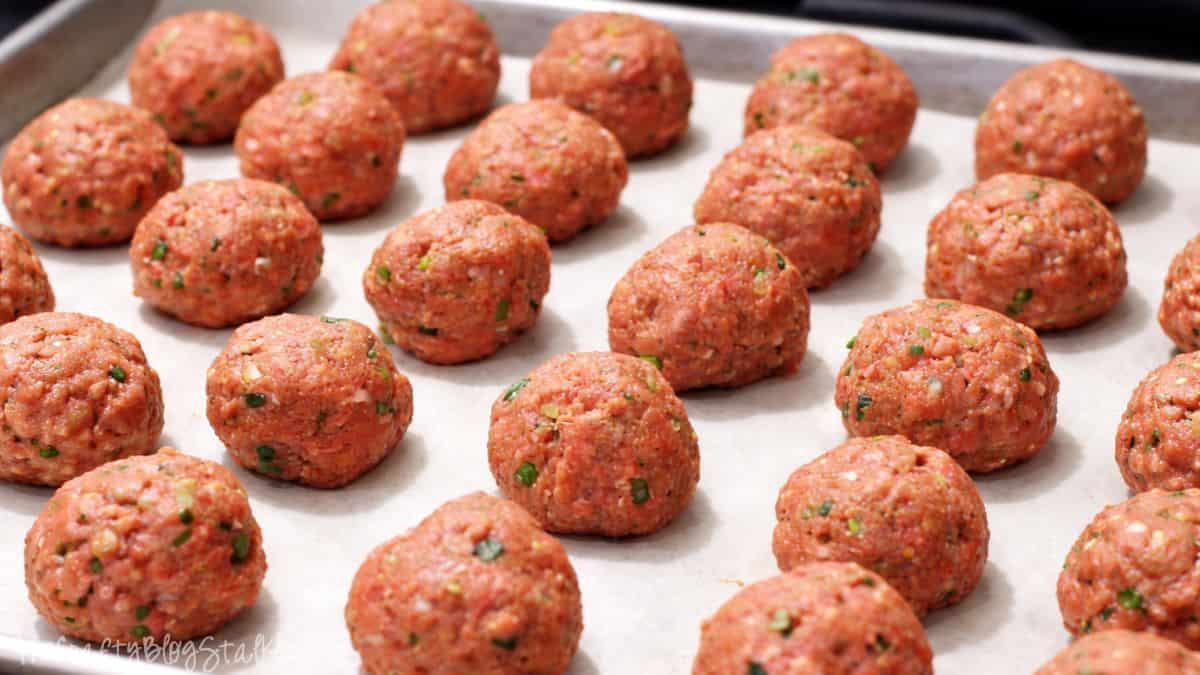 Raw meatballs arranged on a parchment-lined baking sheet, ready to be baked, showing evenly sized meatballs with herbs mixed throughout.