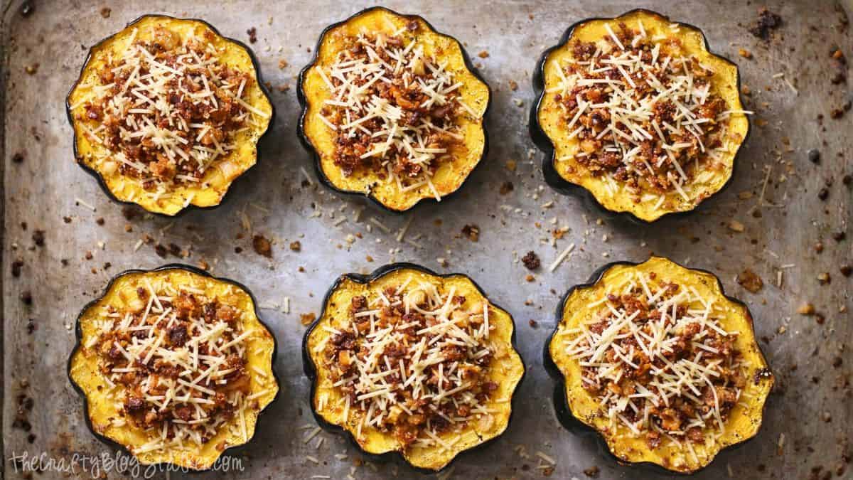 Six halved acorn squash filled with sausage stuffing and shredded cheese, arranged on a baking sheet and ready for roasting.