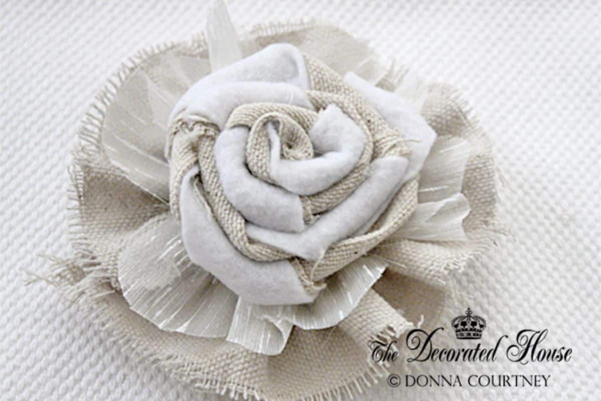Handmade fabric flower made from layered burlap, felt, and sheer material, arranged in a rose shape on a textured background.