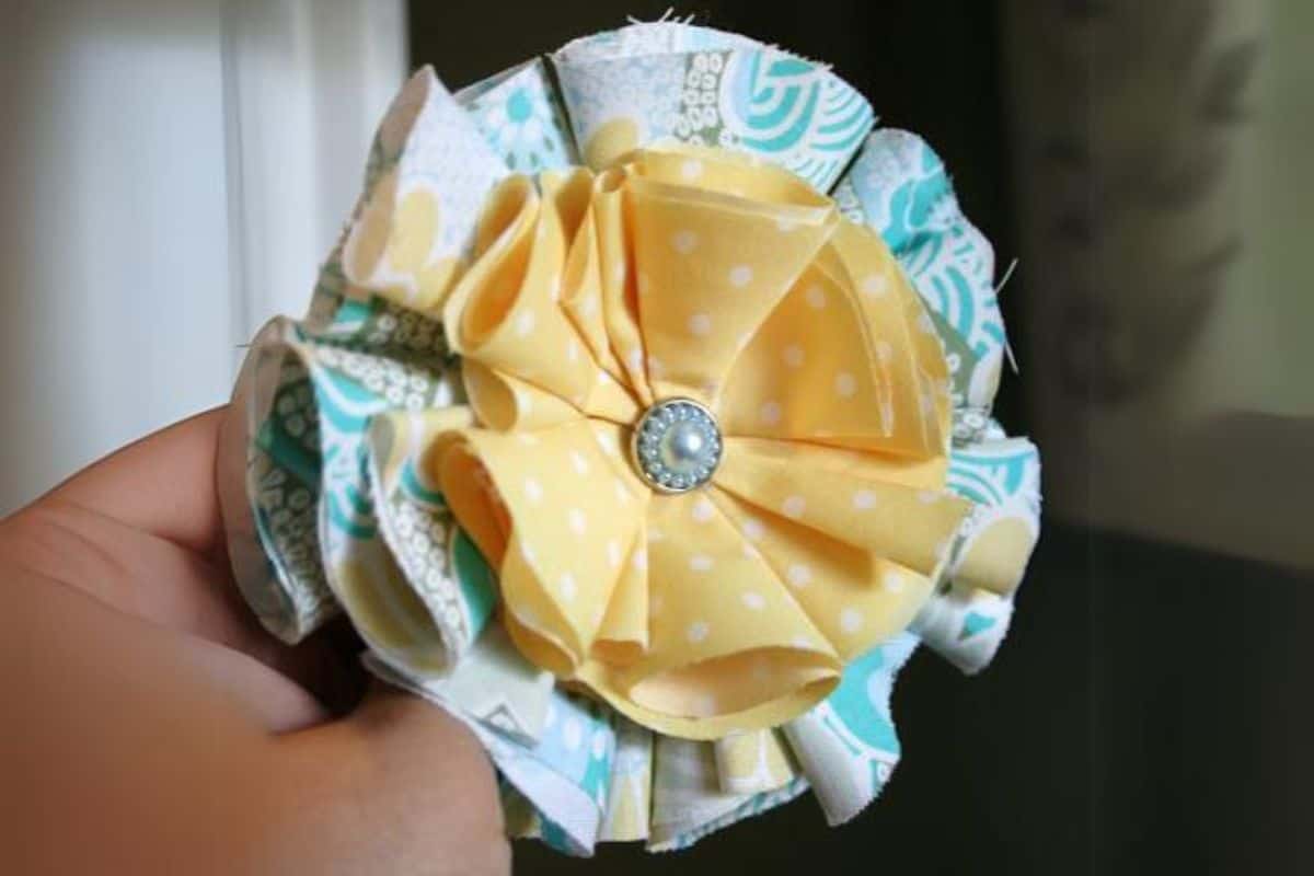Hand holding a layered fabric flower made from folded patterned cotton with a decorative button center, shown close up as a handmade craft.