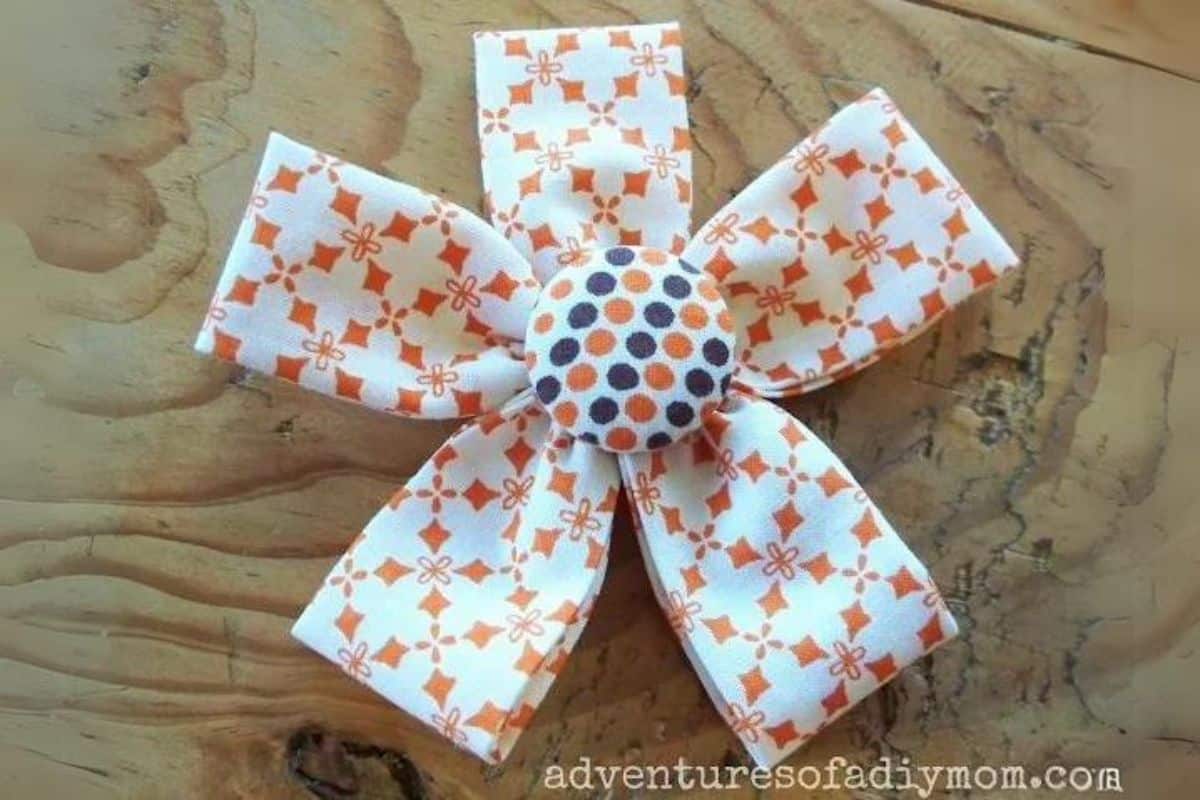 Fabric flower made from folded patterned cotton strips with five petals and a covered button center, displayed on a wooden surface.