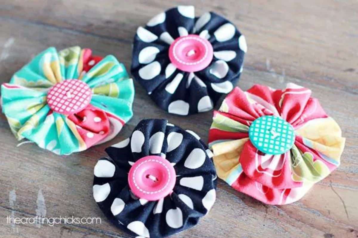 Four handmade yo-yo style fabric flowers with gathered circles and button centers arranged on a wooden surface as DIY craft embellishments.