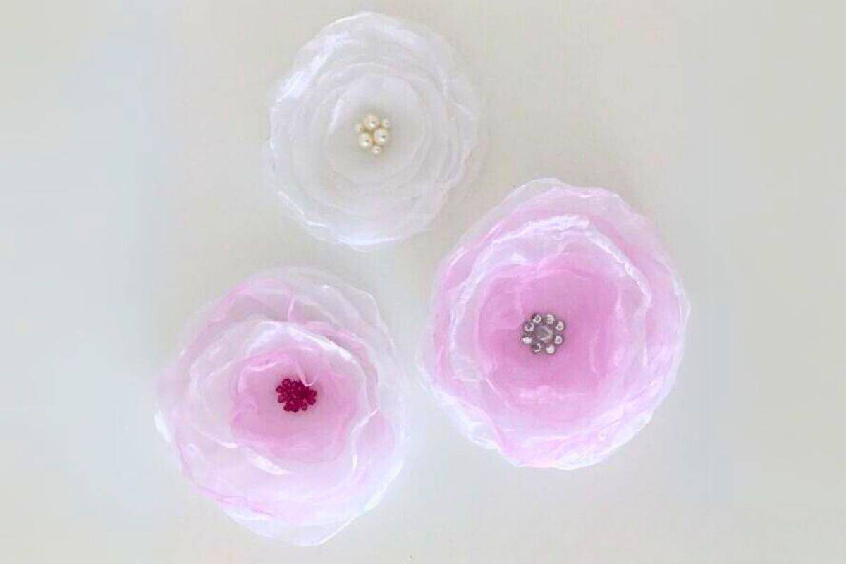 Three handmade organza fabric flowers arranged on a flat surface, each with layered sheer petals and bead centers, used as decorative embellishments.