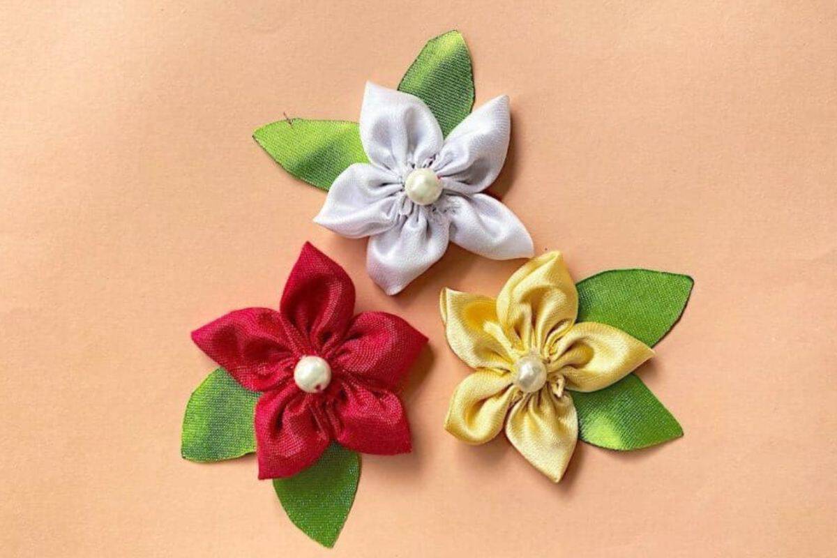 Three handmade satin fabric flowers with layered petals and green leaves arranged on a solid background, each finished with a pearl-style center.