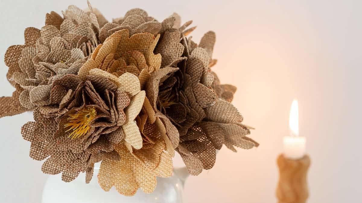 Bouquet of handmade burlap flowers arranged in a glass jar, with layered fabric petals and a lit candle softly blurred in the background.