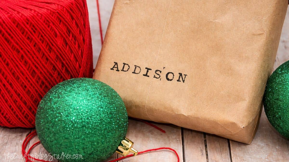 Close-up of a kraft paper wrapped gift stamped with a name, styled with yarn and decorative ornaments on a wooden surface.