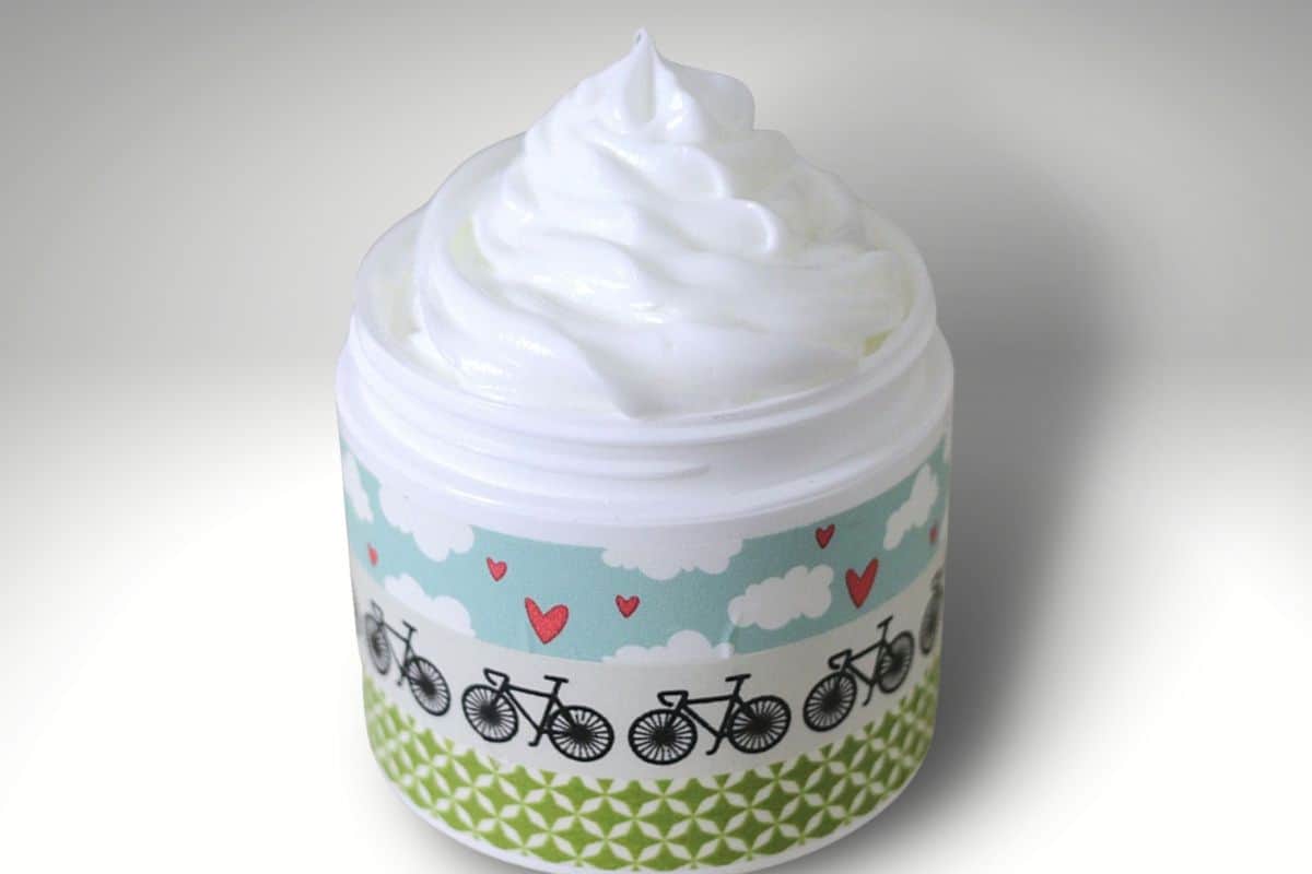 Open jar of homemade whipped lotion with a smooth, fluffy swirl on top and a decorative patterned label, photographed on a light background.