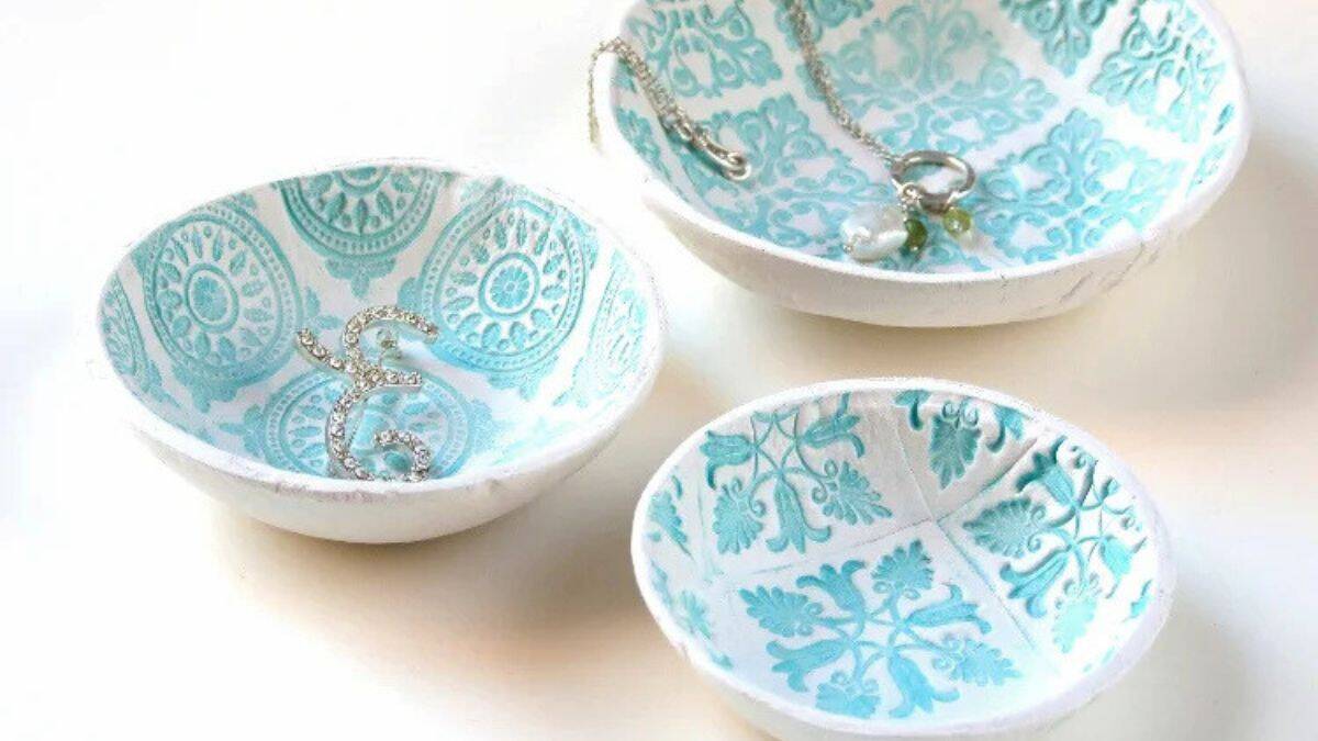 Three handmade stamped air-dry clay bowls with decorative patterns, displayed on a table and holding small jewelry pieces like rings and necklaces.