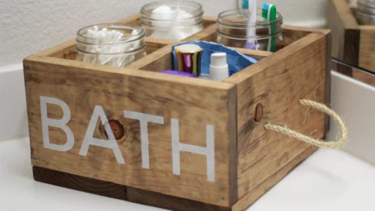 A wooden bathroom caddy with rope handles holding glass jars filled with cotton swabs, cotton balls, toothbrushes, and toiletries on a bathroom counter.