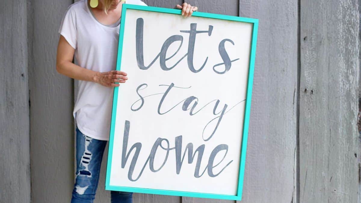 Person holding a large framed wooden sign with a hand-lettered quote, standing in front of a weathered wood wall.
