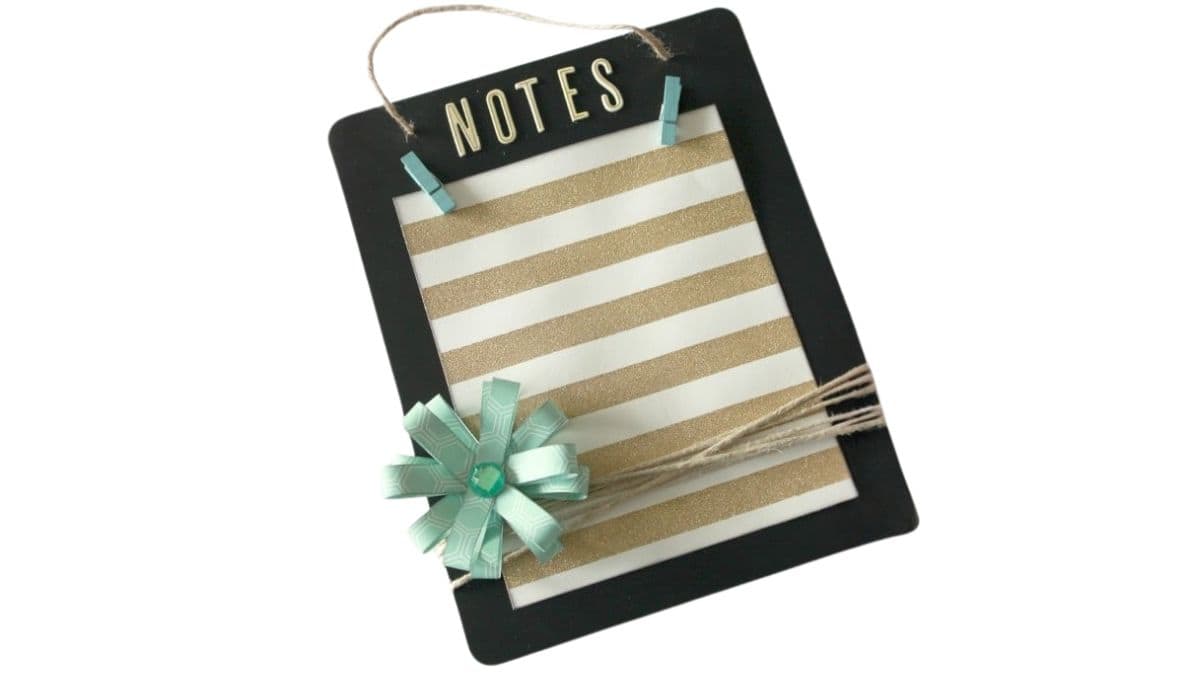 DIY hanging notes board with a small clipboard frame, striped paper, mini clothespins, twine wrap, and a decorative paper flower accent.