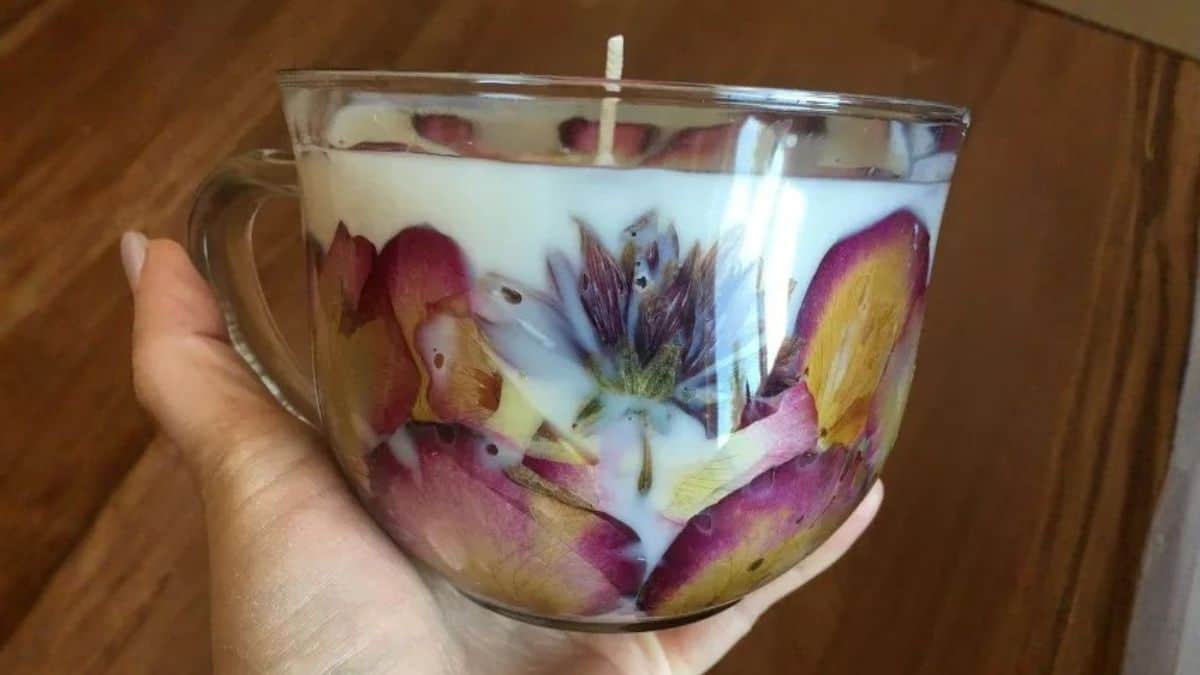 Hand holding a clear glass mug candle made with pressed flowers embedded in the wax and a centered wick.