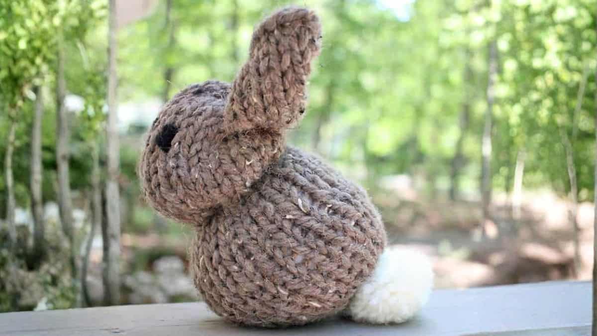 Hand-knitted stuffed bunny viewed from the back, with floppy ears and a fluffy tail, sitting on a wooden surface outdoors with trees in the background.