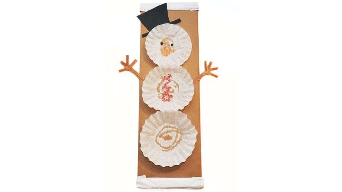 Finished coffee filter snowman craft made with stacked coffee filters, paper hat and nose, and pipe cleaner arms, attached to a brown paper gift bag.