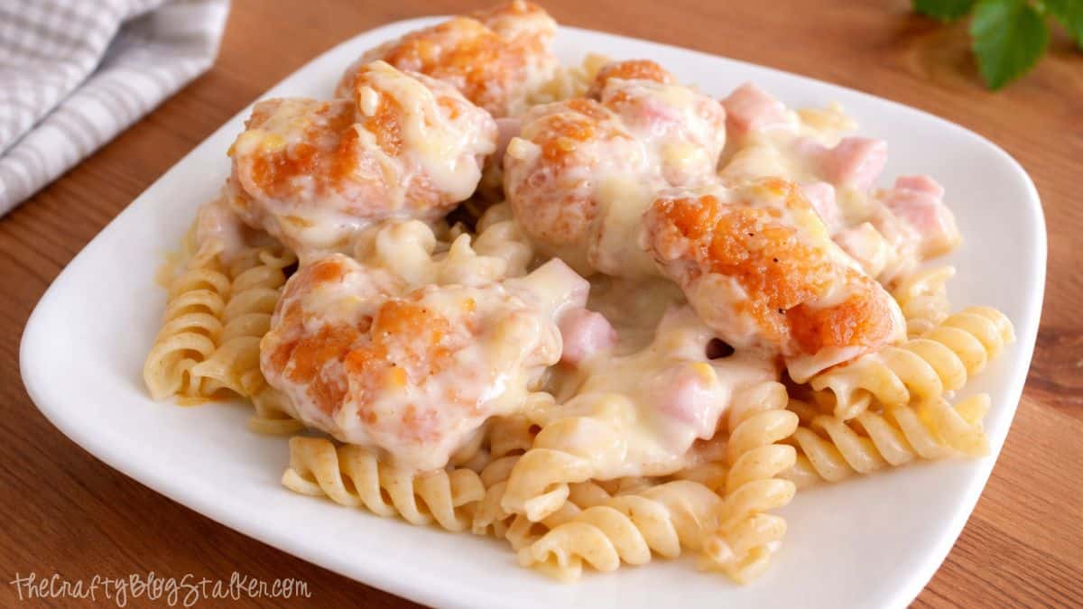 Chicken cordon bleu casserole served over rotini pasta, topped with crispy popcorn chicken, diced ham, and creamy melted cheese sauce on a plate.