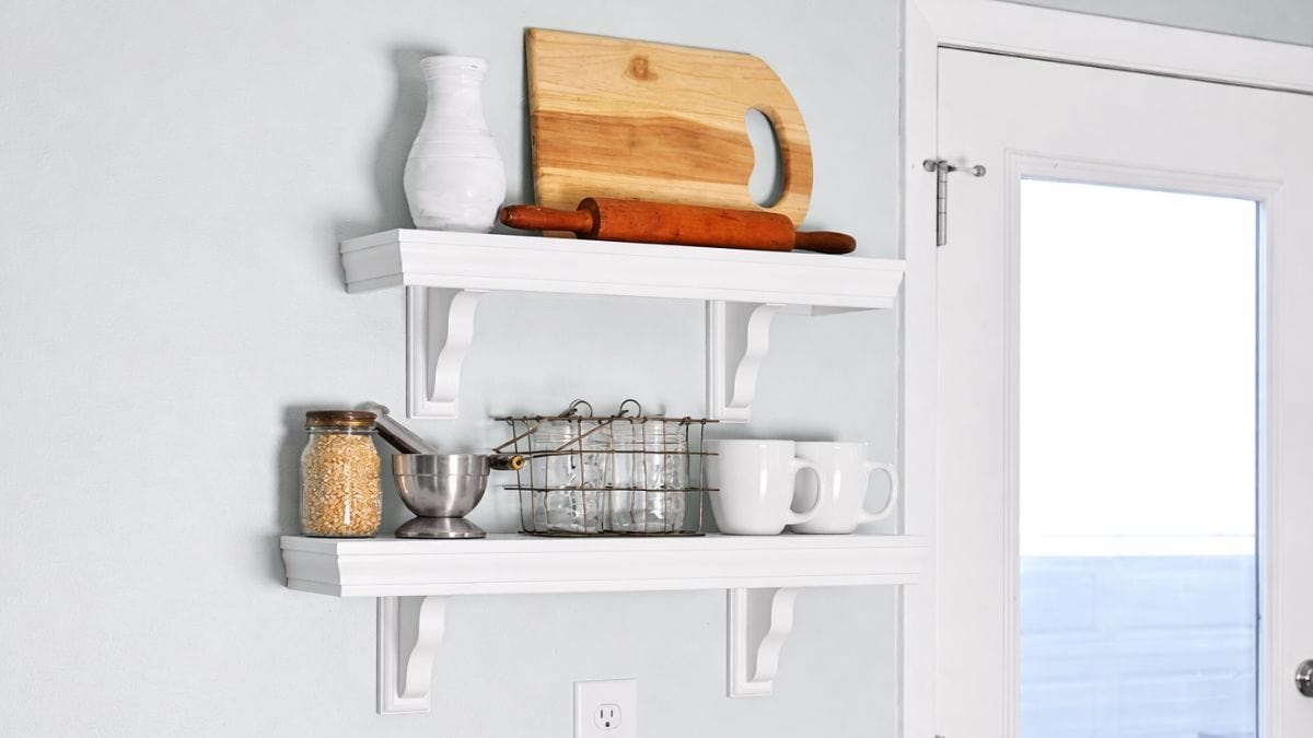 White wall shelves styled with a cutting board, rolling pin, mugs, jars, and kitchen items in a farmhouse kitchen next to a glass door.