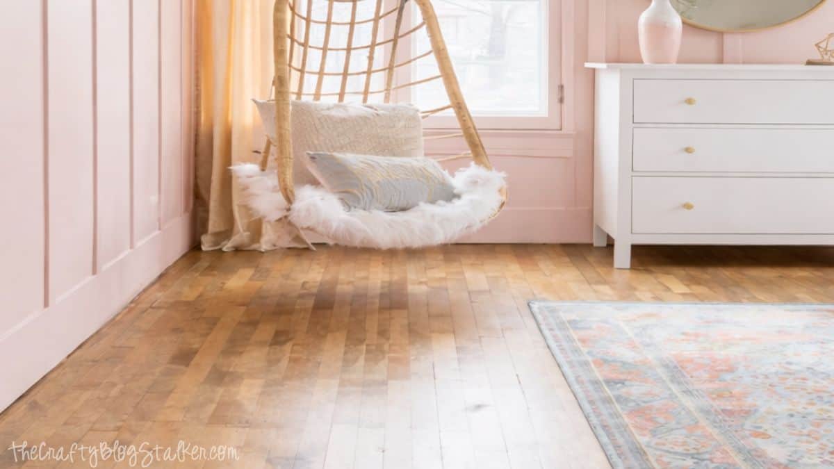 Hanging wicker chair with faux fur seat and pillows near a window, next to a white dresser on hardwood floors in a styled girls bedroom.