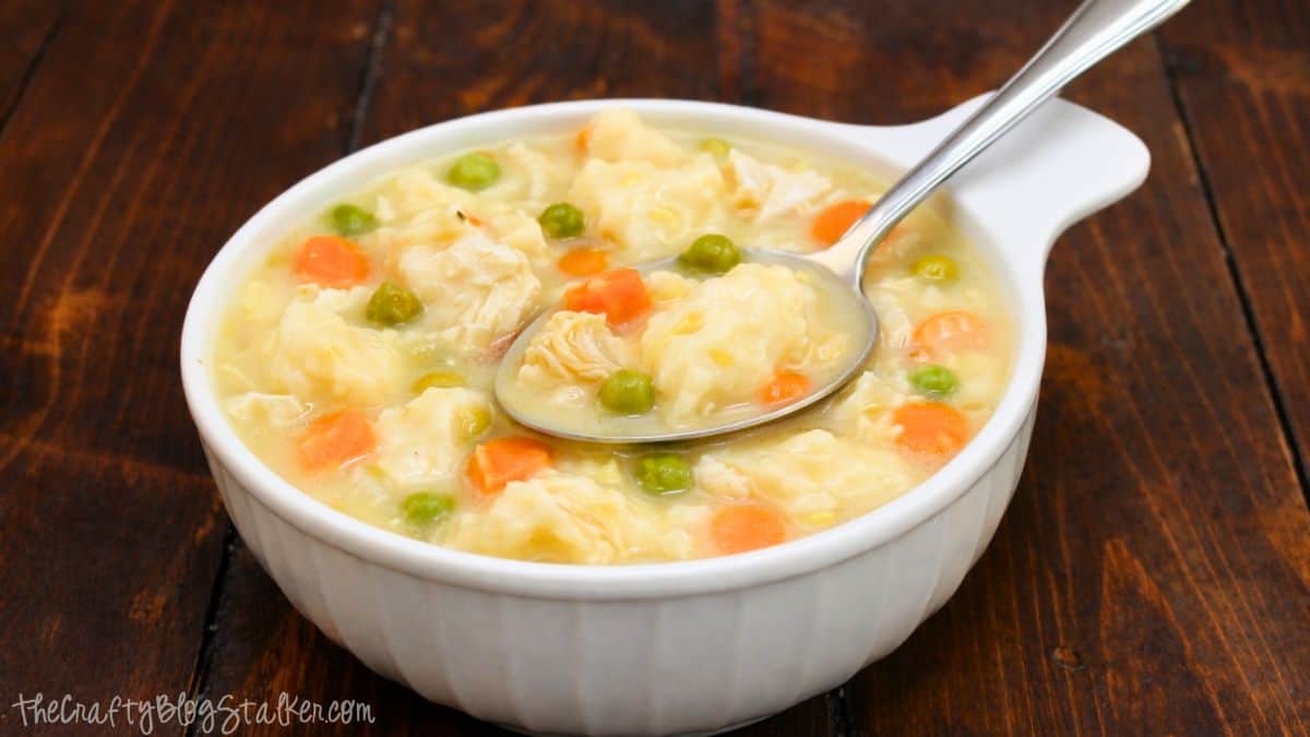 An easy Chicken and Dumplings Recipe to get you ready for the fall! Homemade chicken and dumplings in a white bowl with tender chicken, fluffy dumplings, peas, and carrots in a creamy broth on a wooden table