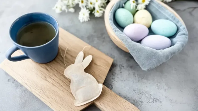 Bunny tea bags made from coffee filters next to a blue cup on a wooden board, next to a basket with Easter eggs