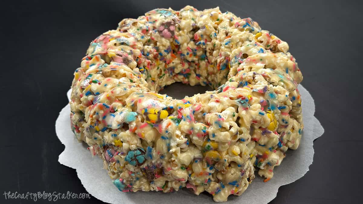 Ring-shaped popcorn cake made from marshmallow-coated popcorn with candy pieces and sprinkles, displayed on parchment paper against a dark background.