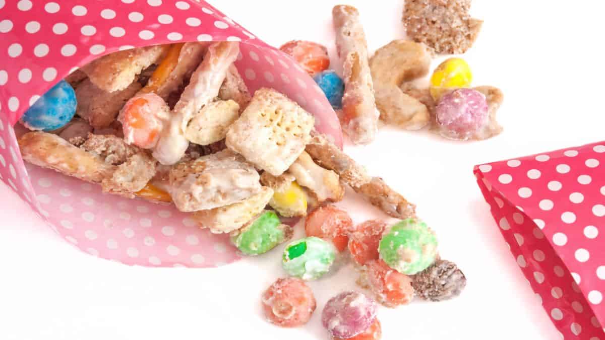 M&M White Chocolate Party Mix Recipe White chocolate party mix with cereal, pretzels, and candy spilling from pink polka dot paper cones onto a white surface.