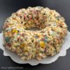 Ring-shaped popcorn cake made with marshmallow-coated popcorn and candy sprinkles, formed like a bundt cake and set on parchment paper.