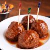 Sweet chili meatball appetizers on a white plate, each topped with a toothpick and coated in a sticky sauce, with extra picks blurred in the background.