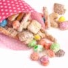 White chocolate party mix spilling from a polka dot paper cone, with pretzels, cereal pieces, and candy-coated chocolates scattered on a white background.
